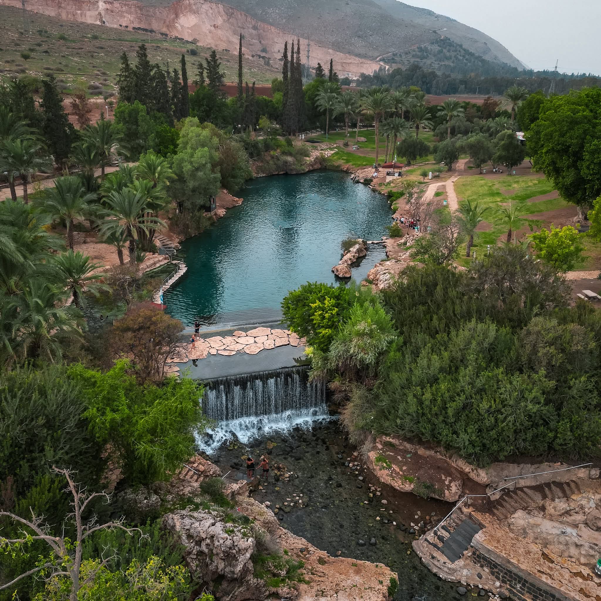 Gan HaShlosha (Sachne) — aerial view of the turquoise spring pool with waterfall and date palms, Mount Gilboa in the background