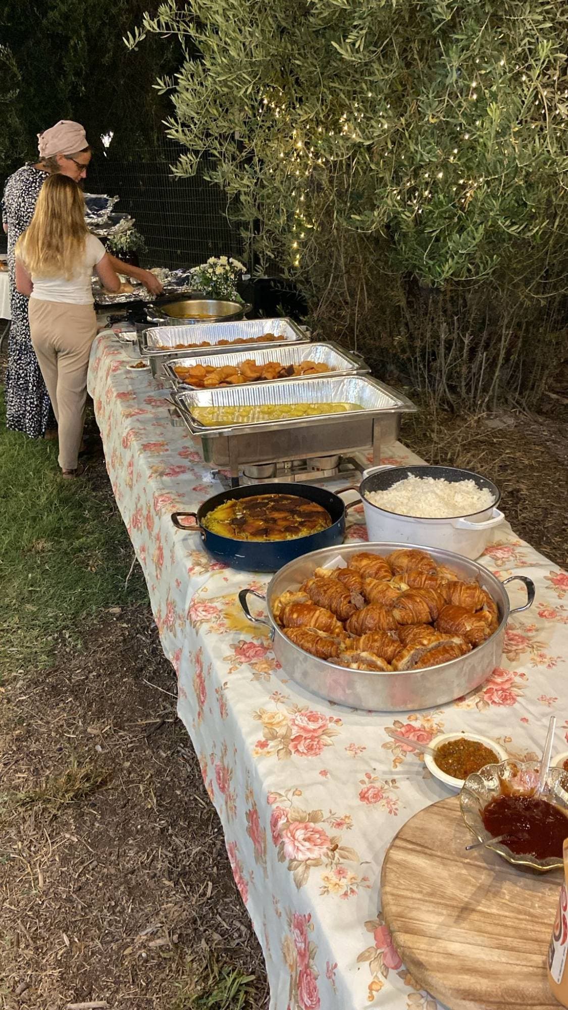 A Bishulilim off-site catering evening — a long buffet table with hot chafing dishes, saffron rice, croissants and salads under olive trees with string lights