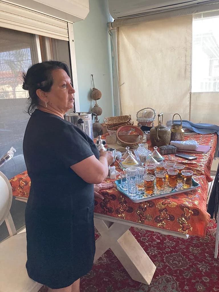 Lili Twizer beside a full Persian tea service — embossed copper teapot, a tray of amber tea glasses on an embroidered orange runner