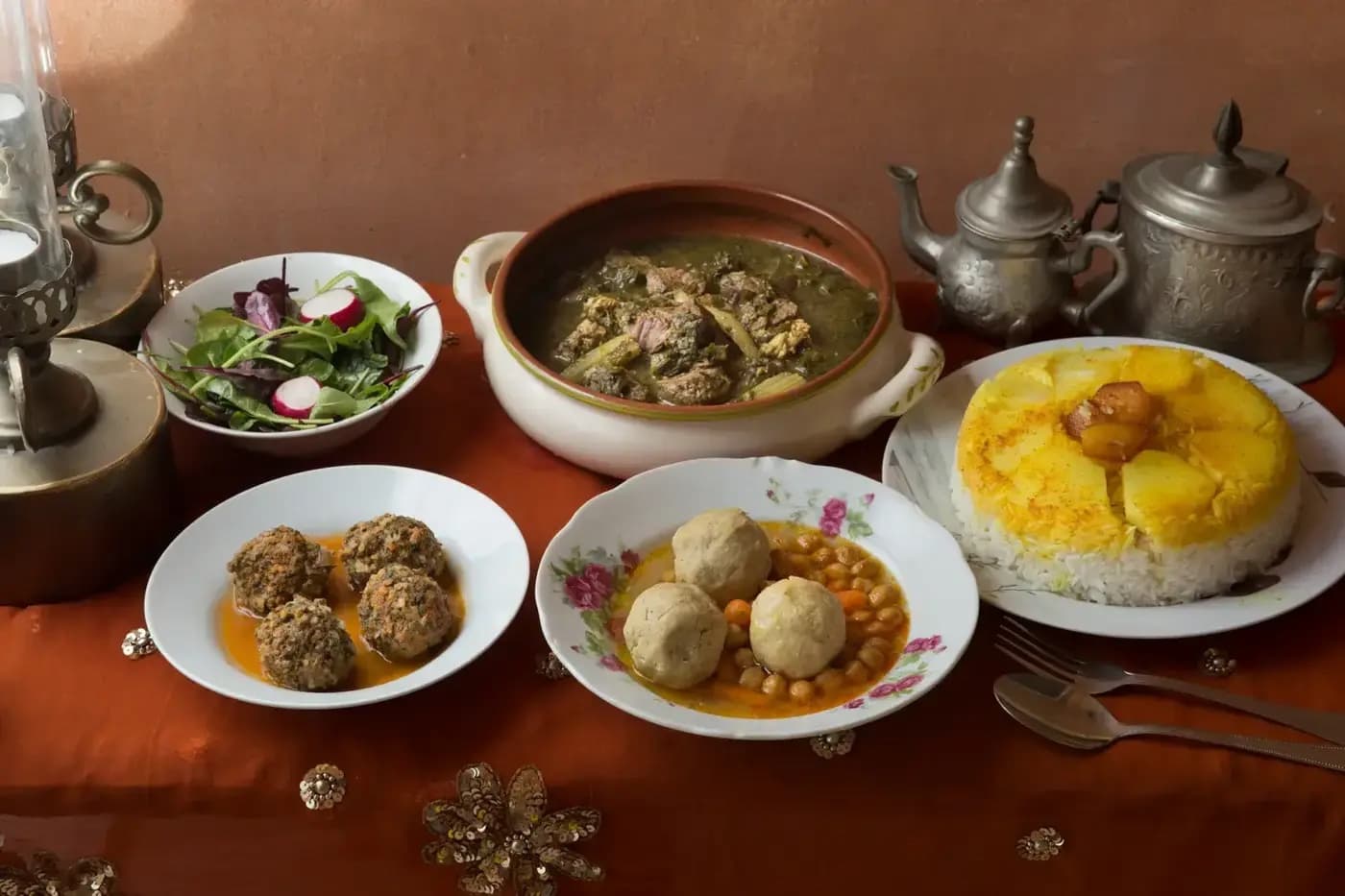 A styled Persian-Jewish table at Bishulilim — Khoresht Sabzi in a ceramic tureen, kotlet, Gondi in a floral bowl, polo with potato tahdig, fresh salad, and Persian silver teapots