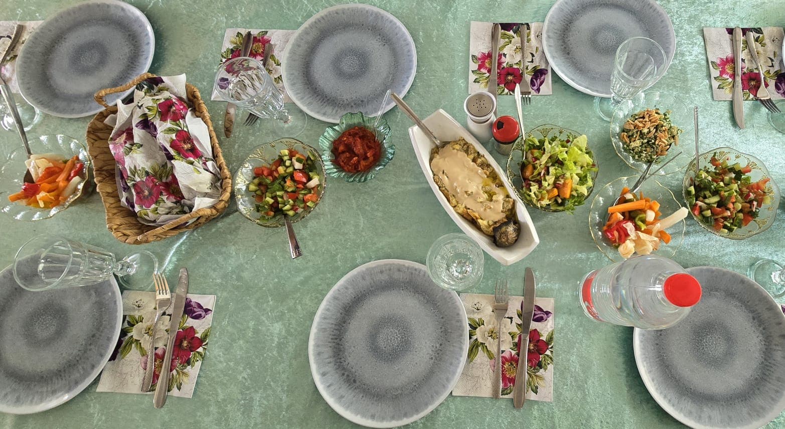 Overhead view of a long table set for dinner — grey plates, floral napkins, salads in glass bowls and a bread basket